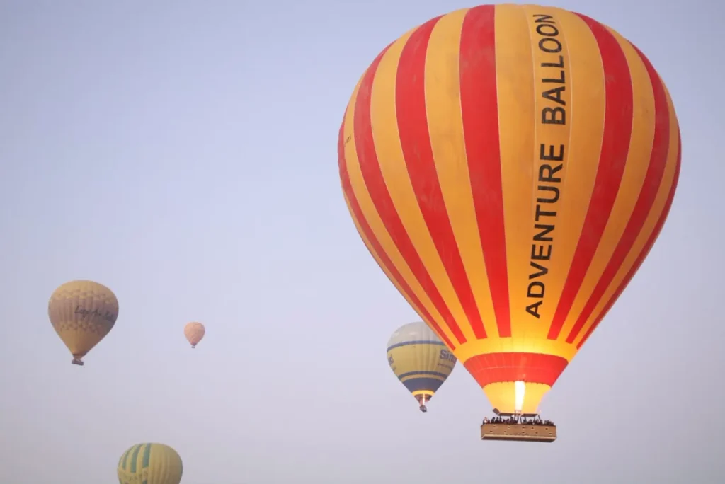 The red and yellow Adventure Balloon envelope flying over Luxor at sunrise, with other hot air balloons in the morning haze