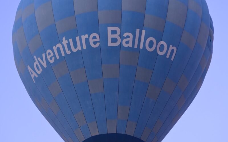 A deep blue hot air balloon marked Adventure Balloon climbing into a clear sky over Luxor