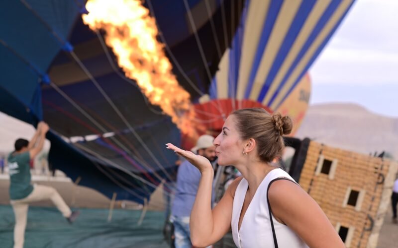 A guest watching a tall burner flame light the inside of an inflating balloon at dawn in Luxor