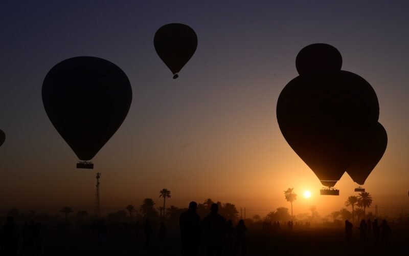 Three hot air balloons silhouetted against a golden sunrise above a line of palm trees in Luxor